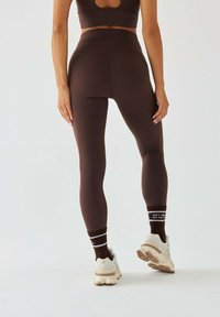 Brązowe legginsy z wysokim stanem o gładkiej fakturze, zestawione z białymi i brązowymi trampkami oraz brązowymi skarpetkami w paski.