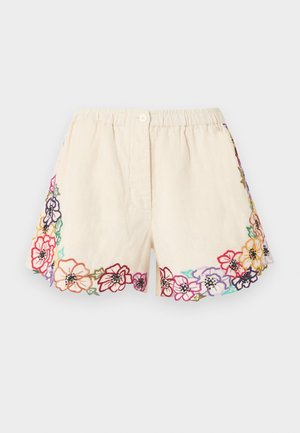 Mujer con blusa color crema y pantalones cortos bordados con flores, combinados con zapatos Mary Jane marrón oscuro, posando frente a un fondo blanco.