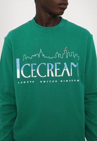 Grøn sweatshirt lavet af bomuld, med et bybillede og ordet "ICECREAM" i fed blå og hvide bogstaver med "LONDON UNITED KINGDOM."