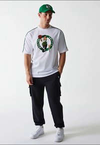 Jersey de malla blanca de manga corta con gráfico de los Boston Celtics, pantalones cargo negros y zapatillas blancas. Gorra verde con el logo de los Celtics complementa el atuendo.