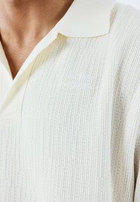 Polo shirt blanc en tissu texturé avec un motif côtelé, doté d'un logo brodé subtil. Col classique, manches courtes, tissu léger.