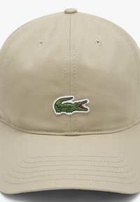Bež bejzbolska kapa od izdržljive tkanine, s zelenim logotipom krokodila izvezenim sprijeda. Zakrivljeni vizir s teksturiranom površinom.
