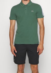 Man wearing green short-sleeve polo shirt with logo on chest and black shorts standing against a plain background.