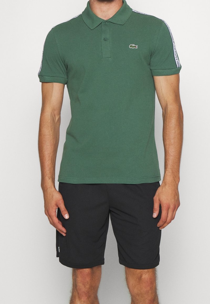 Man wearing green short-sleeve polo shirt with logo on chest and black shorts standing against a plain background.