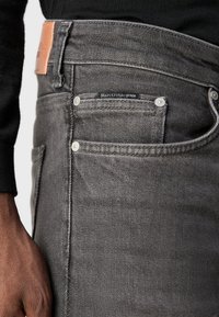 Graue Jeans aus Denim mit einer Nahaufnahme des Bundes, die ein Lederpatch, silberfarbene Hardwareakzente und kontrastierende Steppdetails zeigt.