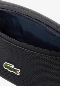 Pochette en tissu noir texturé avec fermeture éclair partiellement ouverte montrant l'intérieur bleu foncé et un logo crocodile vert brodé sur le devant.