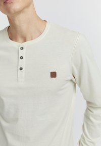 Camisa de manga larga de color beige claro con cuello redondeado, tapeta de tres botones y un pequeño parche de logo marrón en el pecho. Textura suave.