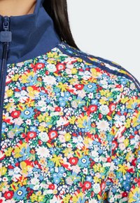 Blommig zip-jacka med ett flerfärgat mönster på en blå bakgrund, med hög krage och broderad logotyp.