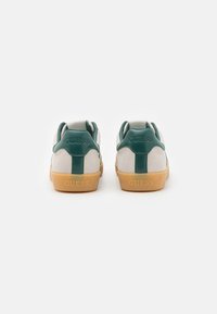 Baskets blanches avec des accents verts et une texture en daim atténuée. Elles présentent une semelle en caoutchouc et un logo « GUESS » sur le talon. Design simple.