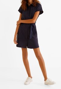 Robe bleu marine boutonnée à manches courtes avec une ceinture nouée à la taille, portée avec des baskets blanches, présentée en position debout.