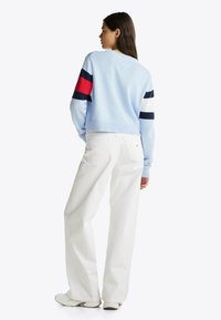 Jasnoniebieski krótki sweter z granatowymi i czerwonymi paskami na rękawach, zestawiony z białymi spodniami o szerokich nogawkach i jasnym obuwiem, widoczny od tyłu.