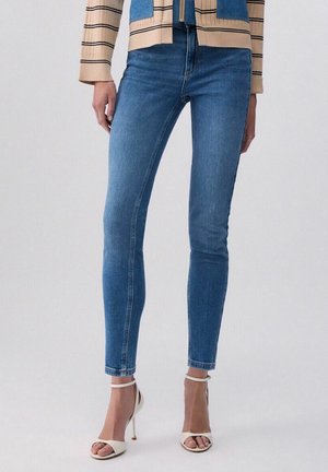 Donna con jeans skinny blu, giacca beige a righe e sandali bianchi con tacco alto, in piedi davanti a uno sfondo semplice.