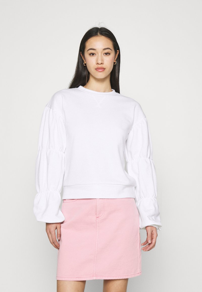 Sweatshirt branco com mangas bufantes e bainha canelada, combinado com uma saia de ganga rosa. A textura parece suave e o design é descontraído.