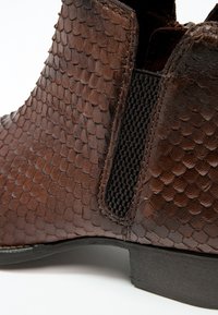 Gros plan d'une botte cheville en cuir marron avec un motif texturé semblable à des écailles, un panneau latéral élastique noir et un petit talon noir.