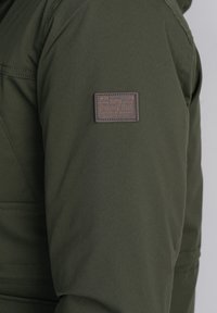 Chaqueta verde oscuro con una etiqueta de cuero marrón en el hombro izquierdo, con un acabado texturizado y sutiles detalles de costura.