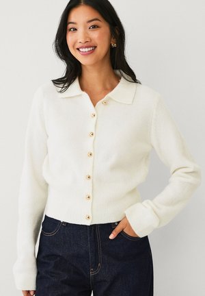 Woman wearing a cream-colored buttoned cardigan with gold buttons and dark blue jeans, smiling with one hand in pocket.