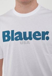 T-shirt bianco in cotone con bold blu con la scritta "Blauer." e "USA" in carattere grigio più piccolo. Collo rotondo semplice e maniche corte.