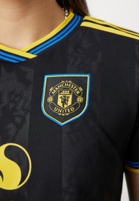 Camisa preta do Manchester United com um tecido texturizado, detalhes em amarelo e azul, e um emblema bordado do clube no peito.