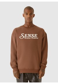 Brun sweatshirt med rund halsringning, ribbade ärmar och tryckt text fram som säger "SENSE Blazing Horizon." Mjuk textur, lös passform.