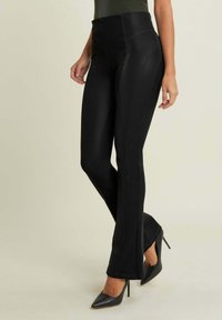 Pantaloni neri a vita alta con un design aderente, texture liscia, dettagli delle cuciture visibili e una leggera svasatura all'orlo, abbinati a tacchi neri.