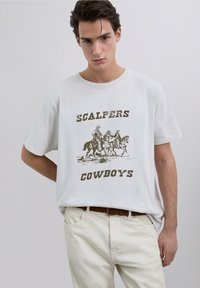Biała bawełniana koszulka z grafiką przedstawiającą kowbojów na koniach, z napisem "SCALPERS COWBOYS" w odważnych, brązowych literach poniżej wzoru.