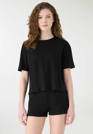 Giovane donna con capelli ricci che indossa una maglietta nera semplice a maniche corte e pantaloncini neri, in piedi davanti a uno sfondo bianco.