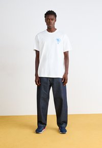 Λευκό βαμβακερό t-shirt με μπλε γραφικό στην αριστερή πλευρά του στήθους, συνδυασμένο με φαρδιά σκούρα τζιν παντελόνια και ναυτικές αθλητικές παπούτσια.