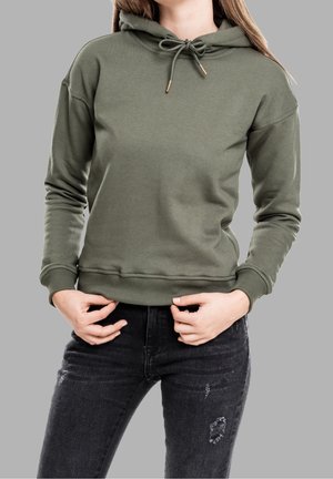 Personne portant un sweat à capuche vert olive et un jean noir déchiré tenant l’ourlet du sweat sur un fond gris uni.