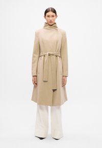 Abrigo de mezcla de lana beige con un alto cuello, cintura ceñida con cinturón y bolsillos laterales. Mangas largas y un corte relajado, complementado por una textura suave.