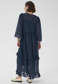 Marineblaues, ärmelloses Maxikleid mit weißen Polka-Dots, ausgestattet mit gestuften Rüschen am Saum und lockerem Sitz. Leichtes, transparentes Material.