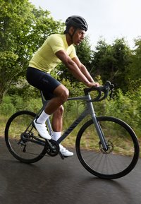 Vélo gris de canyon avec des roues noires, cycliste portant un tranche de jaune vif et un short noir, chaussures blanches, roulant sur une route mouillée avec de la verdure en arrière-plan.