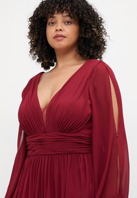 Burgundy dress with a deep V-neck, pleated bodice, and chiffon sleeves. Features a ruched waist and sheer details on the sleeves.