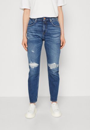 Μπλε τζιν παντελόνι με ψηλή μέση, με ραγισμένα γόνατα και ελαφρώς στενή γραμμή. Συνδυασμένο με λευκά αθλητικά παπούτσια.