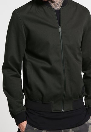 Homme portant une veste bomber vert foncé zippée sur une chemise noire et un pantalon camouflage, les mains dans les poches.