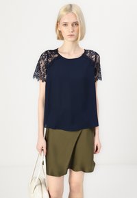 Modra bluzica z čipkastimi kratkimi rokavi in okroglim izrezom, kombinirana z olivno zeleno krilo. Model drži bež torbico.