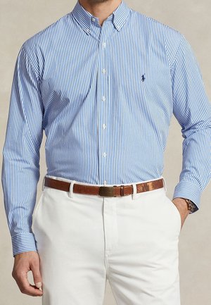 Homme portant une chemise à boutons rayée verticalement bleu clair et blanc, rentrée dans un pantalon blanc avec une ceinture en cuir marron, une main dans la poche.
