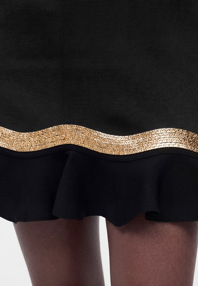 Μαύρη υφασμάτινη φούστα με κυματιστό χρυσό παγιετένιο περιθώριο πάνω από μια φουντωτή εσοχή, παρουσιασμένη σε γυμνά πόδια πάνω σε ανοιχτό φόντο.