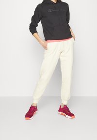 Felpa nera con grande logo, pantaloni jogger crema con polsini elasticizzati e scarpe da ginnastica bordeaux con dettagli arancioni. Il modello è in piedi con una mano sui fianchi.