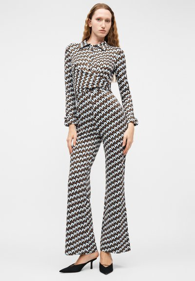 Γυναίκα που στέκεται φορώντας ολόσωμη φόρμα με μπλε και καφέ σχέδια, μακριά μανίκια και φαρδιά πόδια, με μαύρες μυτερές γόβες, μπροστά σε απλό φόντο.