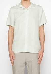 Camicia verde chiaro a maniche corte con bottoni, caratterizzata da un motivo circolare sottile, design con colletto e bottoni bianchi. Indossata sopra pantaloni beige chiaro.