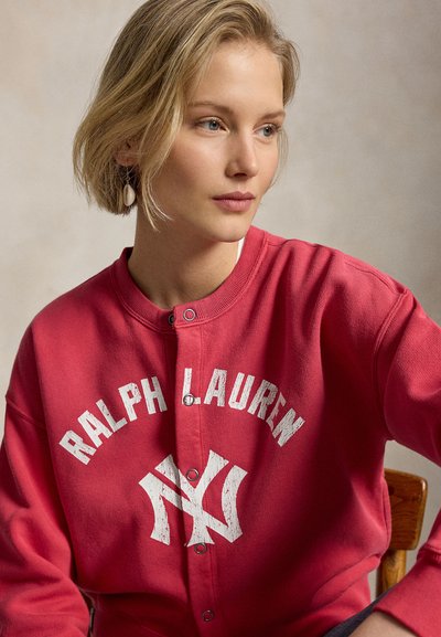 Νεαρή γυναίκα με κοντά ξανθά μαλλιά που φοράει κόκκινο κουμπωτό φούτερ Ralph Lauren και ένα μονό σκουλαρίκι, καθισμένη σε ξύλινη καρέκλα.