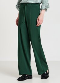 Frau trägt dunkelgrüne weite Hose mit Kordelzug, schwarze Loafers und ein grünes karierte Langarmhemd, das eingesteckt ist.