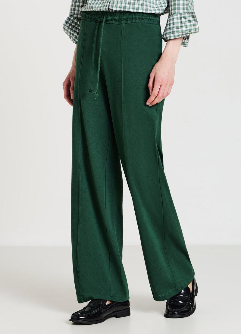 Frau trägt dunkelgrüne weite Hose mit Kordelzug, schwarze Loafers und ein grünes karierte Langarmhemd, das eingesteckt ist.