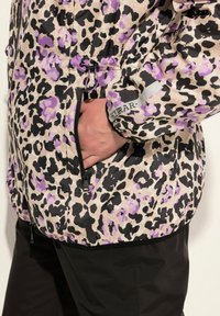 Pälsjacka med leopardmönster i beige, svart och lila nyanser. Har en ficka med dragkedja, justerbar dragsko och elastiska muddar.