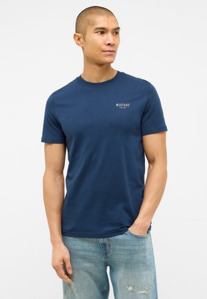 Navy blue cotton t-shirt featuring a crew neck, short sleeves, and a small white logo on the left chest. Paired with distressed light denim jeans.