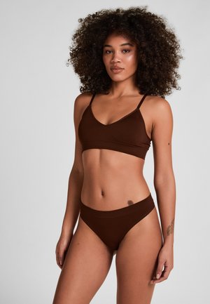 Vrouw met krullend haar, gekleed in een donkerbruine bralette en bijpassend ondergoed, staand tegen een eenvoudige grijze achtergrond.