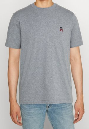 Man draagt een effen grijs T-shirt met korte mouwen, voorzien van een klein rood en blauw logo op de linkerborst, gecombineerd met lichtblauwe spijkerbroek.