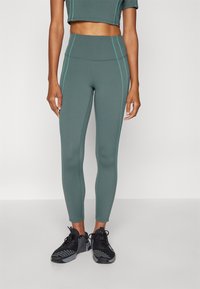 Leggings sportivi verdi con vita alta, caratterizzati da cuciture teal a contrasto. Abbinati a scarpe da corsa nere. Tessuto liscio e elastico.