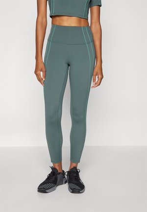 Leggings de sport verts avec une taille haute, dotés de coutures teal contrastantes. Associés à des chaussures de course noires. Tissu lisse et extensible.