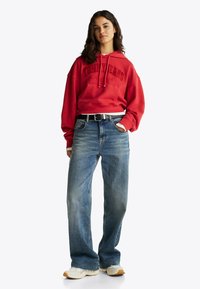 Felpa rossa con cappuccio e tasca frontale, abbinata a jeans a gamba larga blu chiaro. Le scarpe sono crema con accenti beige. Una cintura nera completa il look.